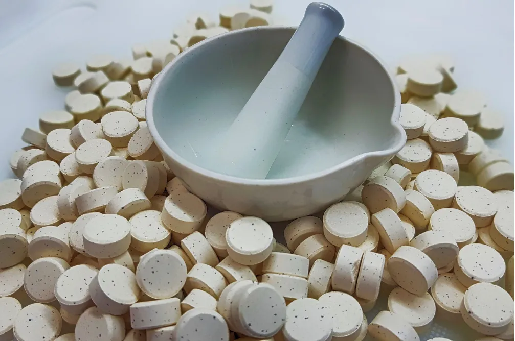A mortar and pestle sits on top of a pile of round, beige tablets or pills.