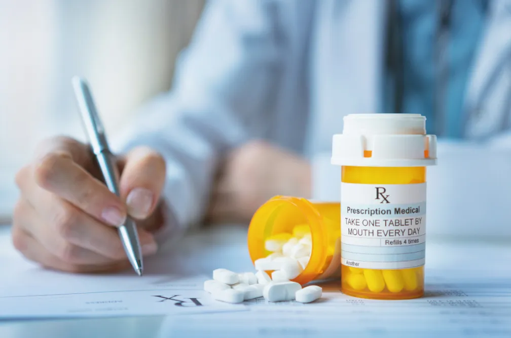 A doctor writes on a clipboard beside an open pill bottle and scattered tablets on a desk.