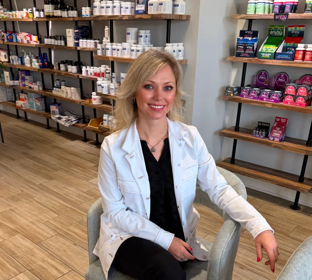 Woman in a white coat sits smiling in a modern pharmacy or wellness store with shelves of products behind her.