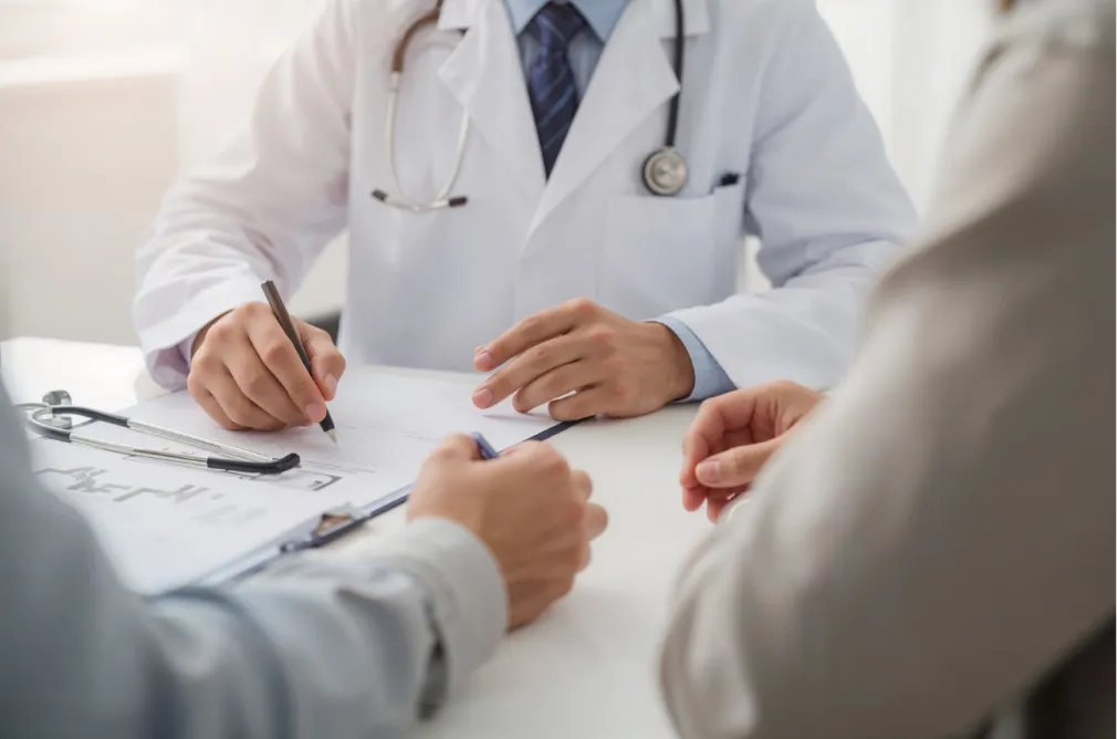 Doctor in white coat discussing a medical form with two patients across a desk in an office.