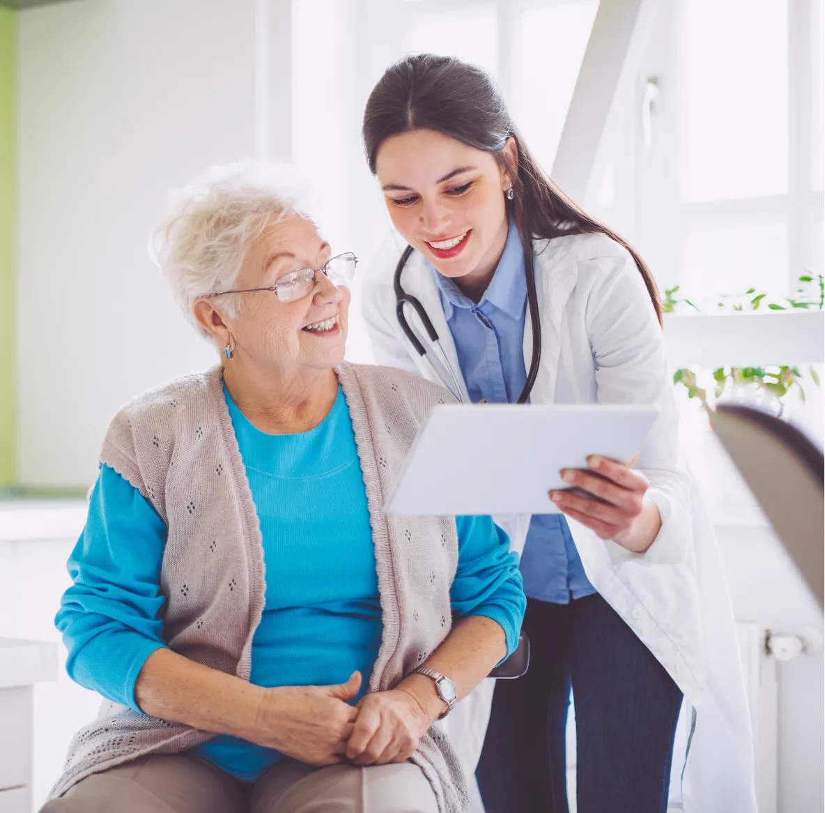 Smiling doctor shows a tablet to an elderly woman sitting and smiling in a bright room.