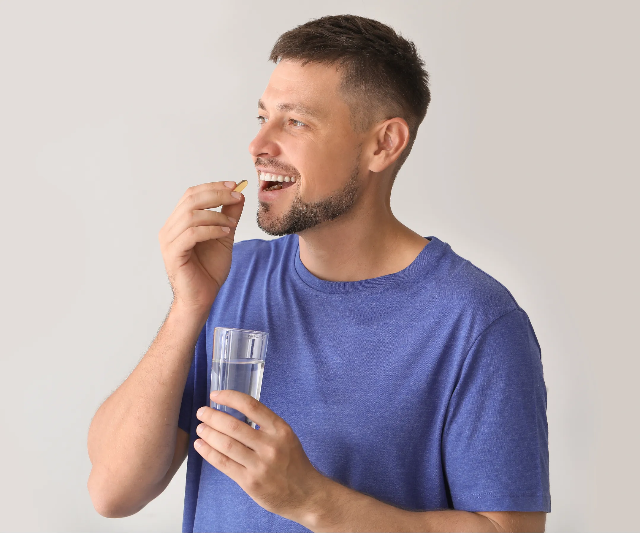 Man in a blue shirt smiling while taking a pill and holding a glass of water.