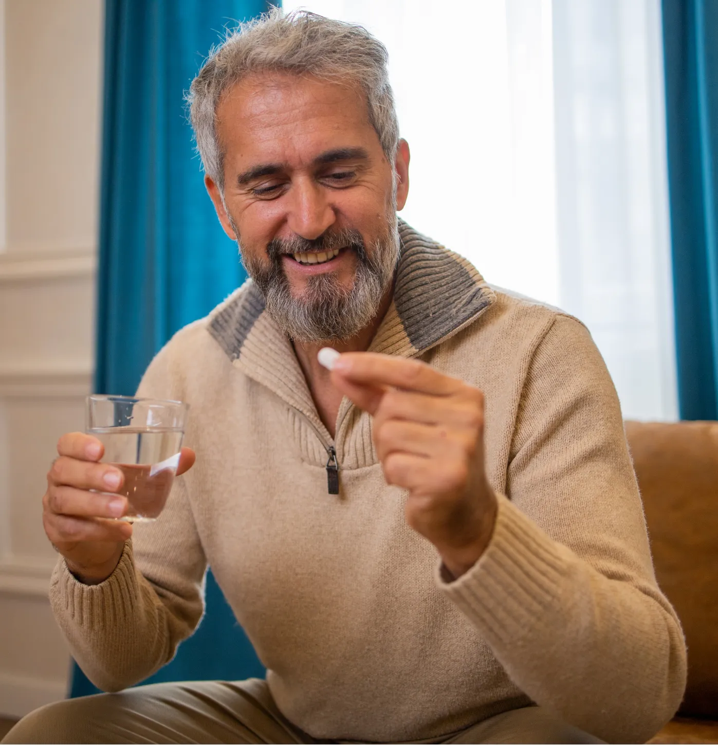 Smiling man holding a glass of water and a pill, sitting indoors on a sofa.
