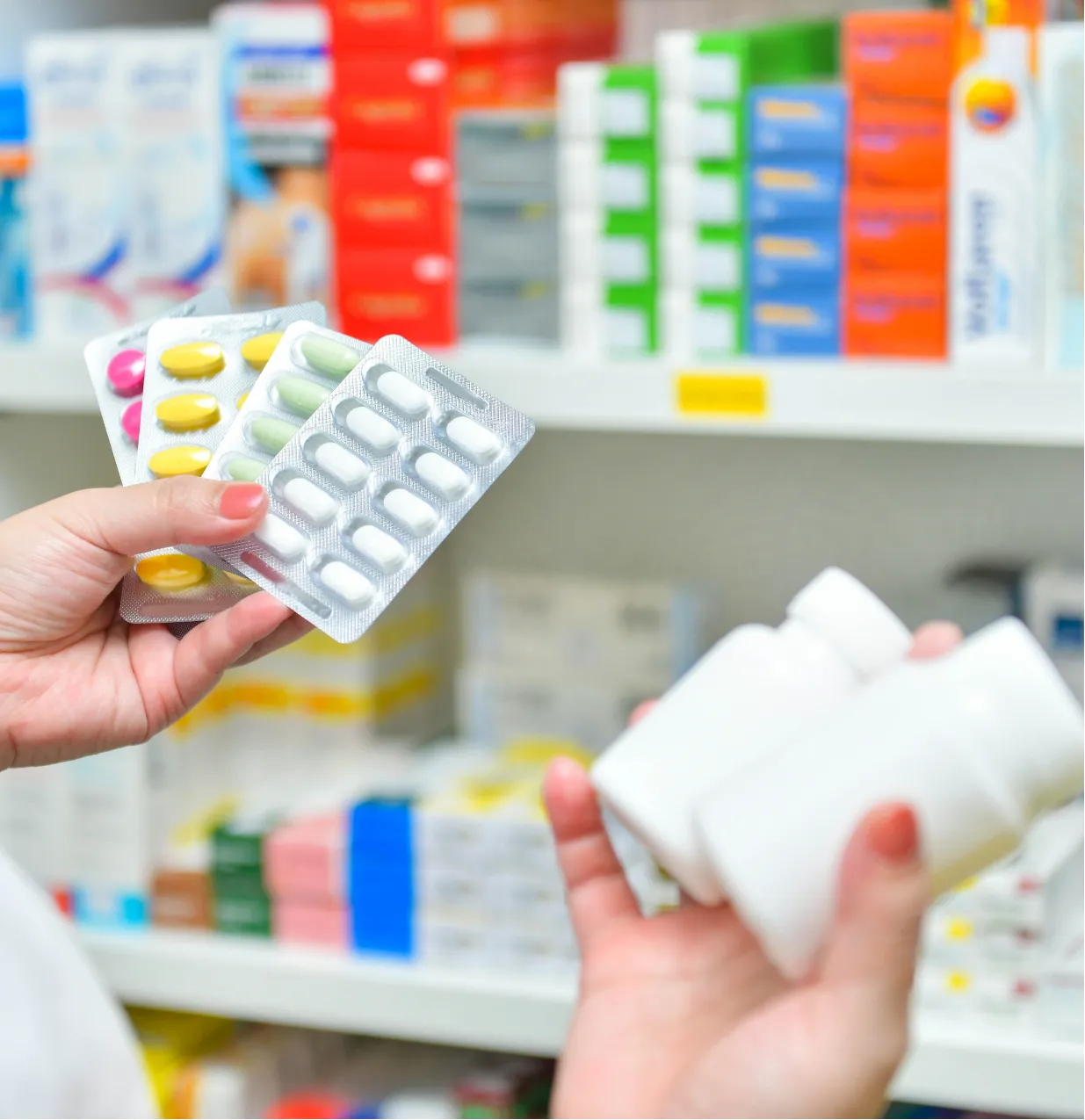 Person holding blister packs of pills and white bottles in a pharmacy with shelves of medications in the background.