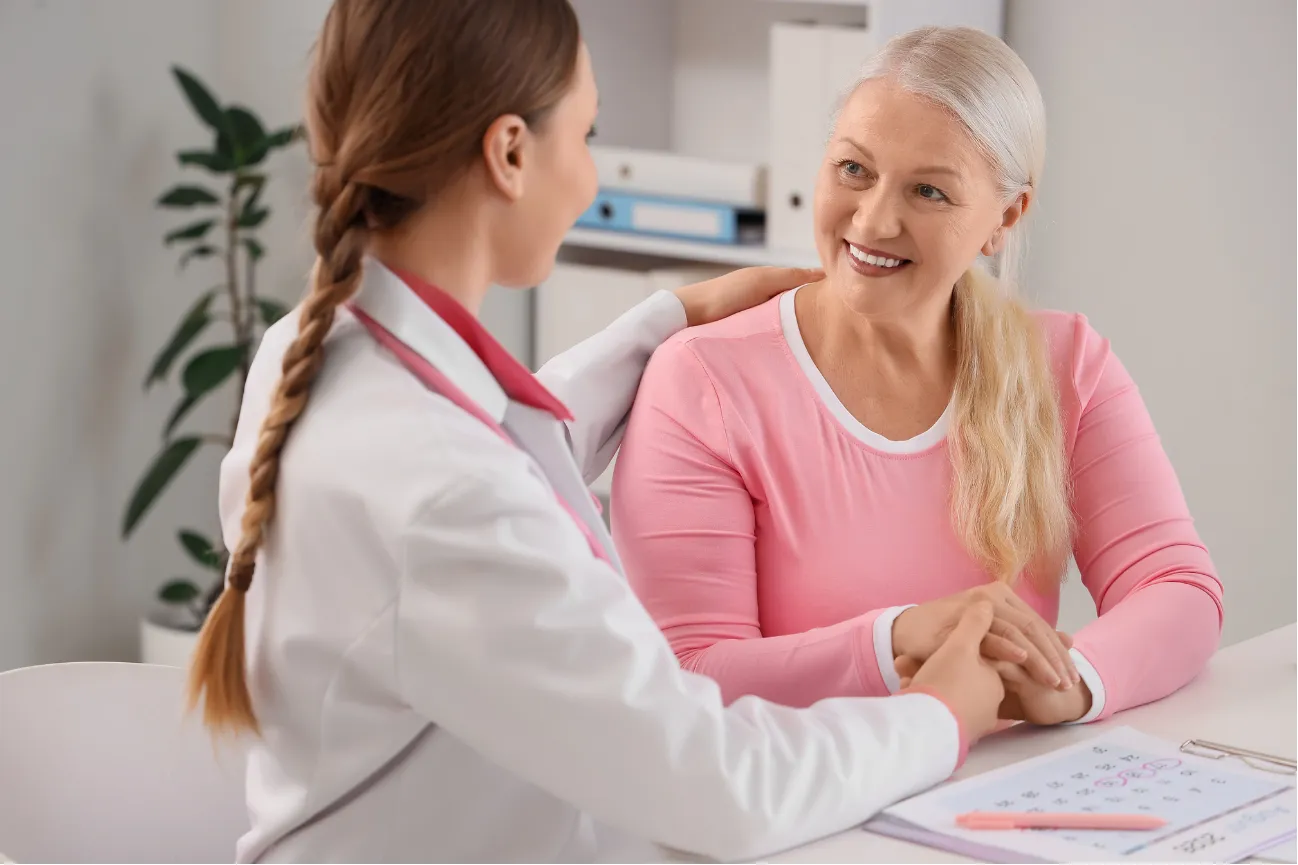 Female doctor comforts smiling older woman during a medical consultation at a bright office desk.
