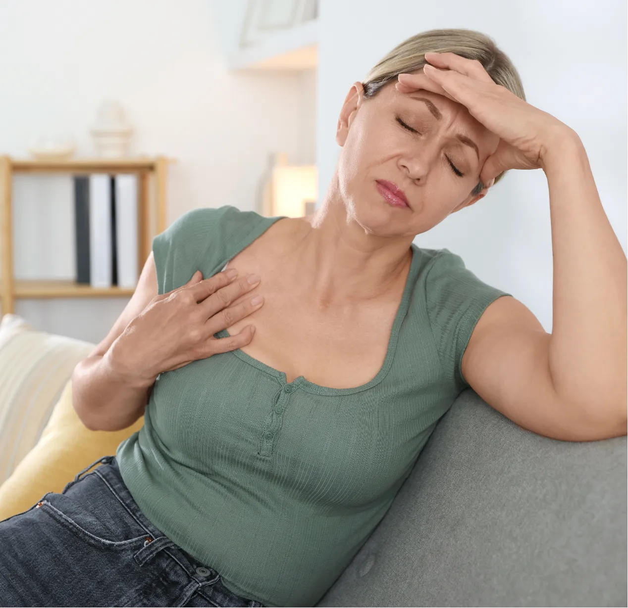 Woman sitting on couch with hand on chest and forehead, appearing to be in pain or discomfort.