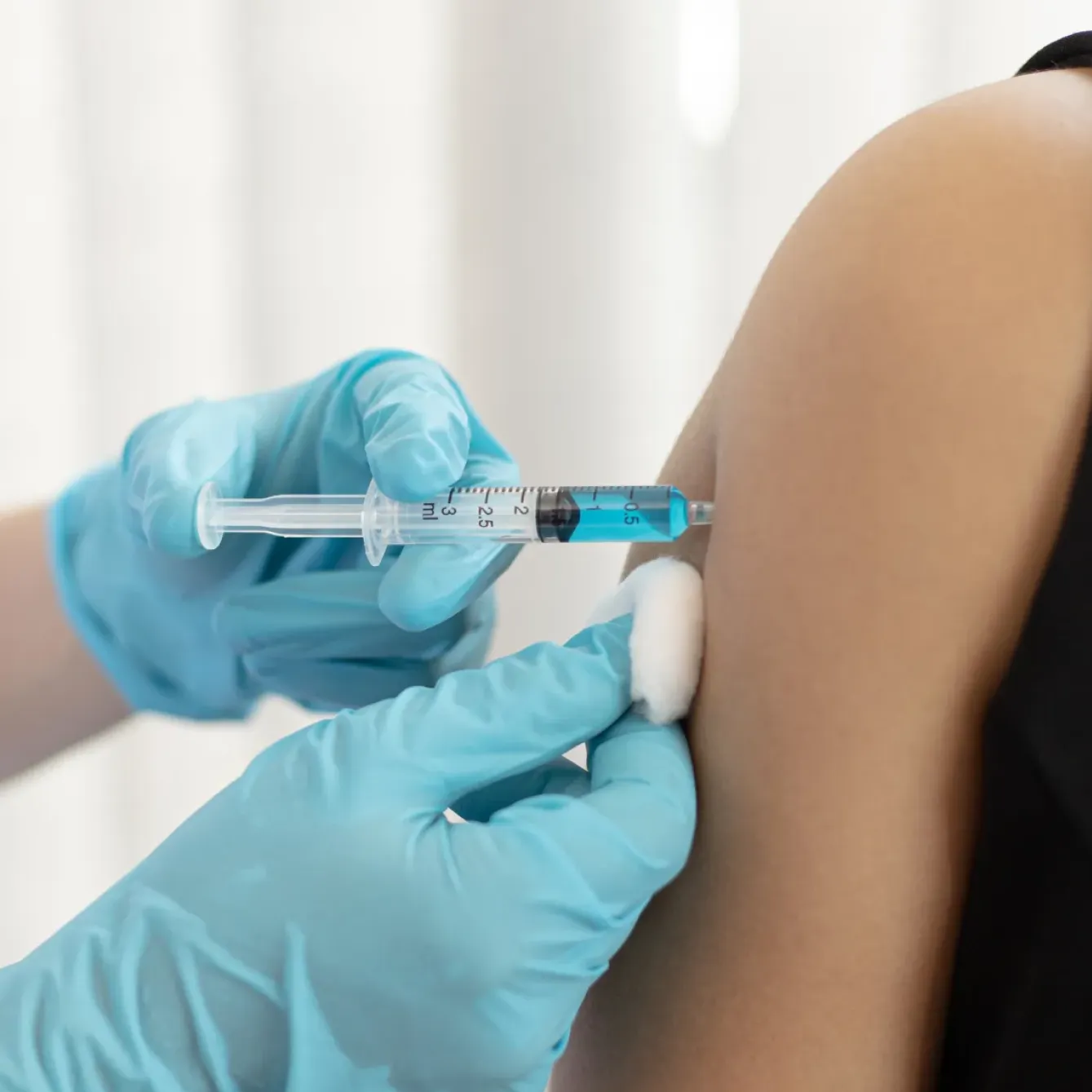 A gloved hand gives an injection with a syringe into a person's arm while holding a cotton swab.
