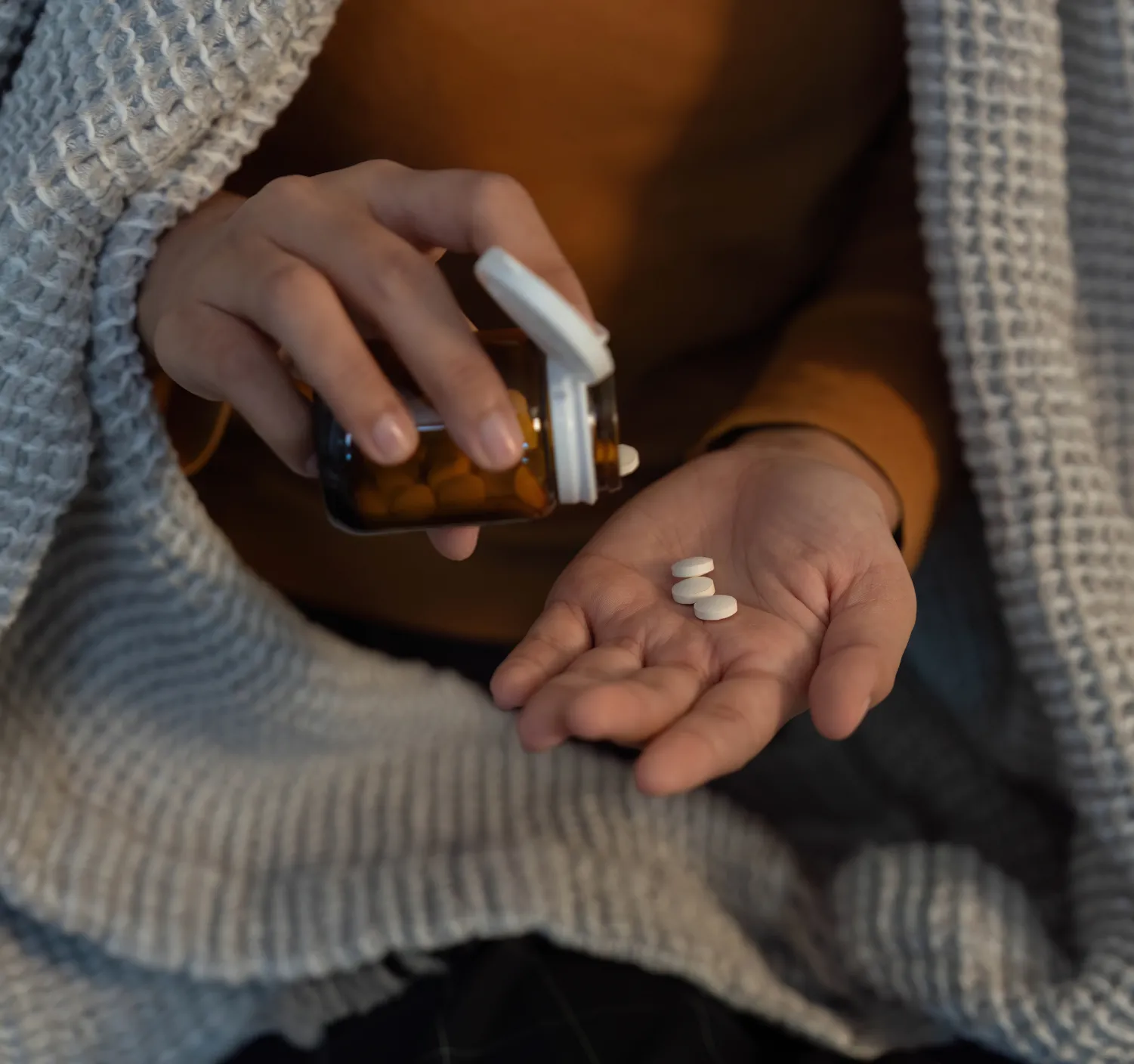 A person pours pills from a brown bottle into their hand while sitting under a gray blanket.
