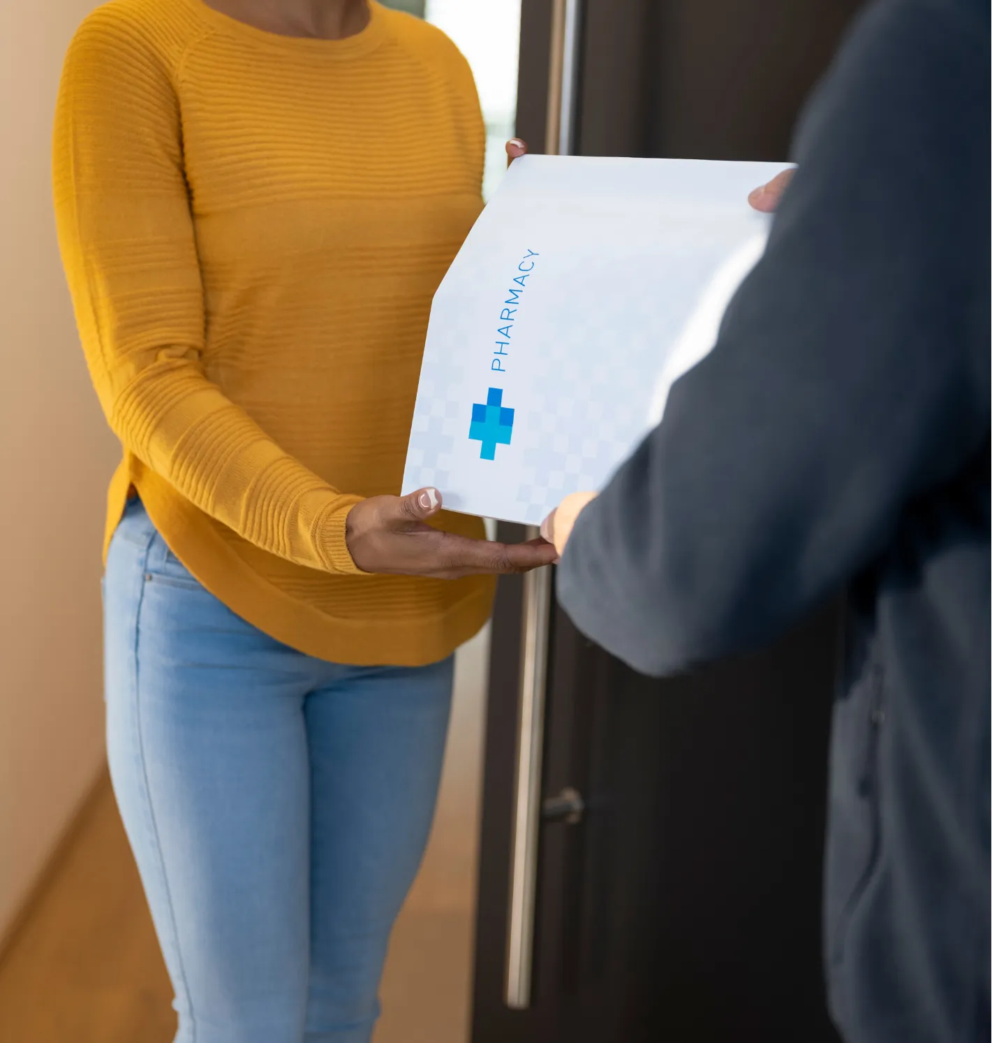 A woman receives a pharmacy package from a delivery person at her door.