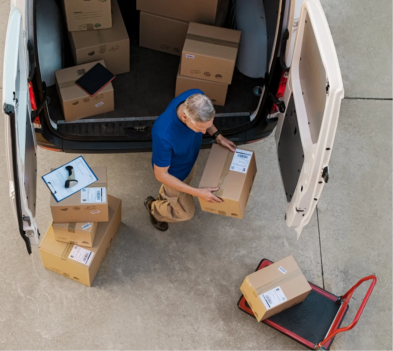 A man unloads cardboard boxes from a van onto the ground and a red dolly in a driveway.