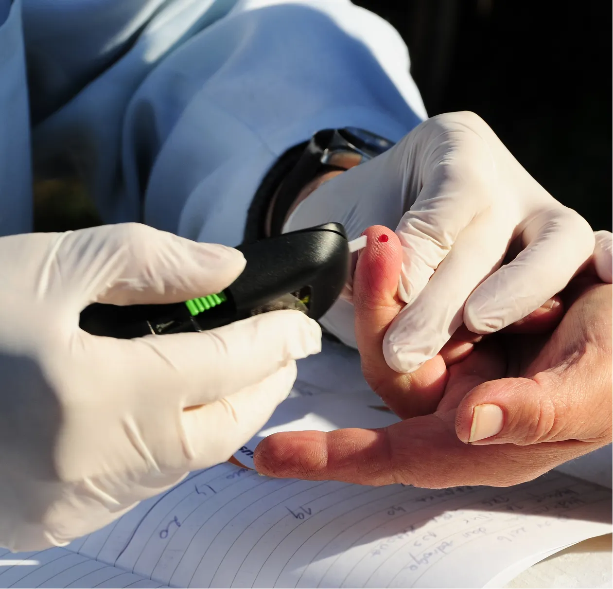 Gloved hands using a glucose meter to test a blood sample from a fingertip over paperwork.