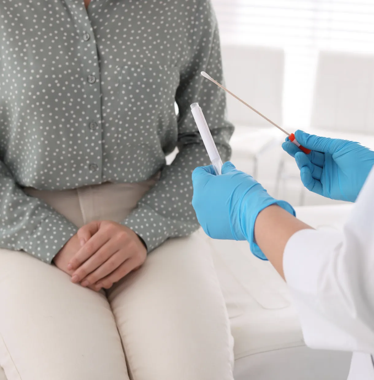 Person in polka dot shirt sitting while a gloved doctor prepares a swab and test tube for a medical test.